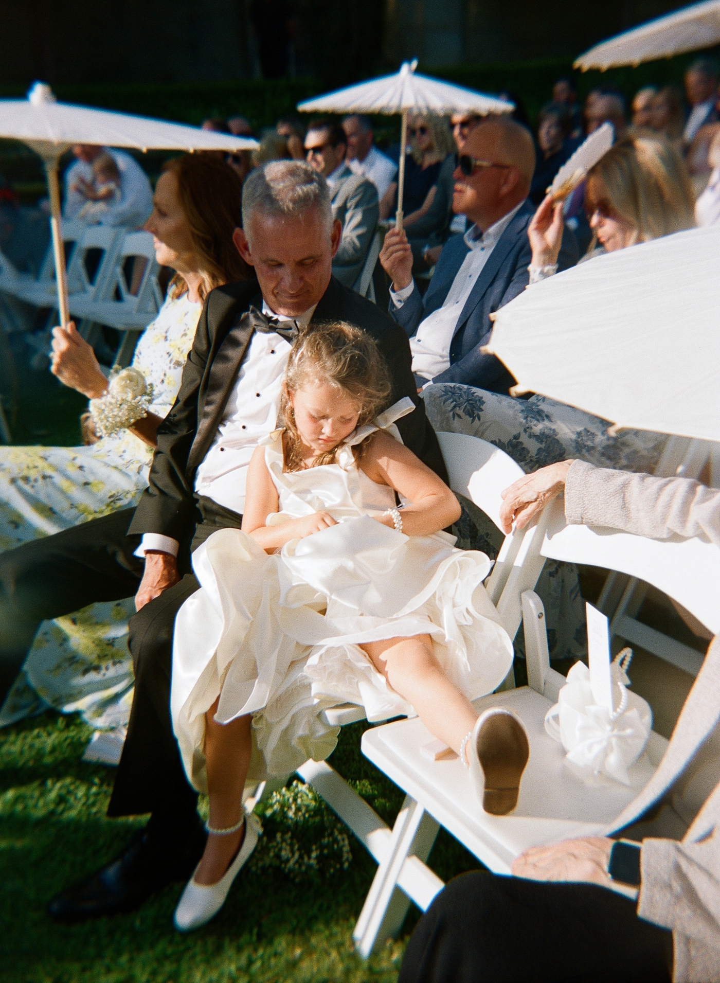 Flower girl lounging across two ceremony chairs and leaning on her grandfather 