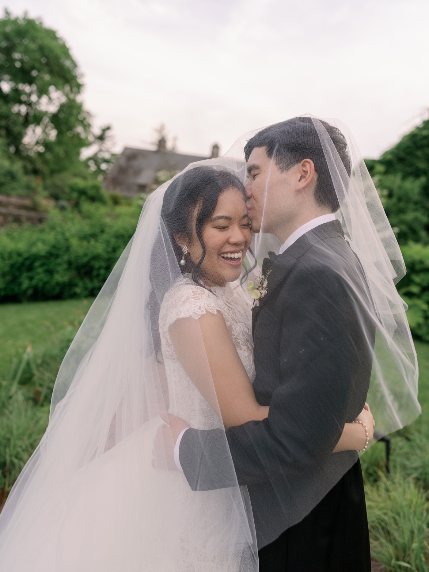 Groom kissing bride on temple while both are draped under her veil 